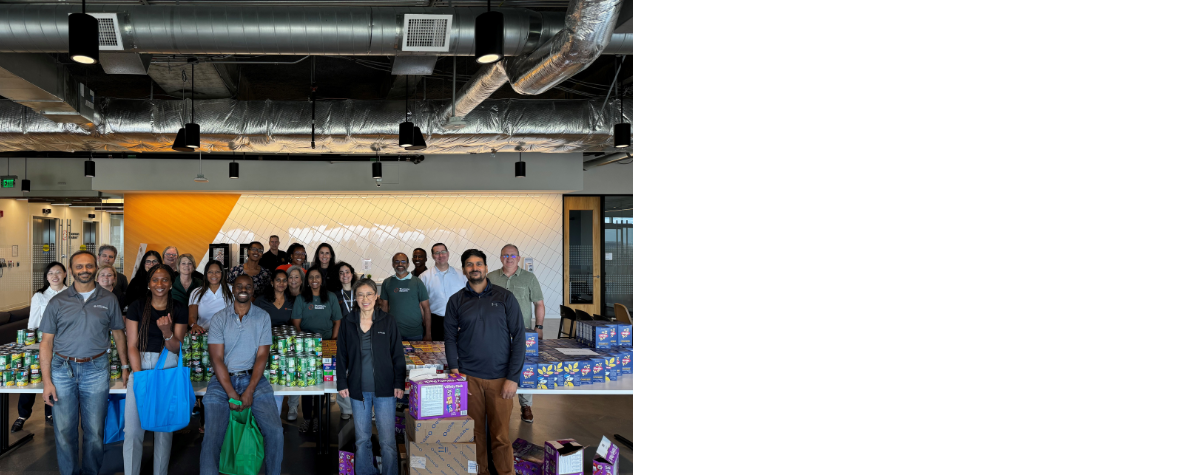 A large group of people stand together during a volunteer event. There are many cans of food on the table around them. 